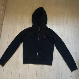 Svart hoodie från Polo Ralph Lauren - Svart hoodie från Polo Ralph Lauren med dragkedja och huva. Klassisk design med röd broderad logga på bröstet och kängurufickor framtill. Tillverkad i mjuk bomull, perfekt för en chill och stilren look.