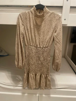 Beige kortklänning med brunt mönster - Supersöt beige kortklänning från Shein med brunt prickigt mönster, hög krage med volang och långa puffärmar. Smockad midja och volangkant nedtill ger en snygg siluett. Perfekt för dig som gillar trendiga detaljer och mjuka material.