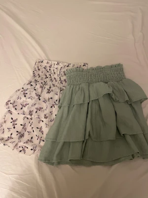 Två volangkjolar  - Säljer två söta korta kjolar. Ena är ljusgrön med flera volanger och smock i midjan, andra är vit med lila och svarta blommor, också med volanger och smock. 1 för 30 eller 2 för 50.