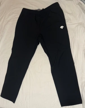 Svarta mjukisbyxor från Descente  - Svarta mjukisbyxor från Descente med vit logga på benet och elastisk midja med snörning. Byxorna har en rak passform och är tillverkade i mjukt syntetmaterial, perfekta för chill eller träning. Snyggt tryck på midjebandet ger extra stilpoäng.