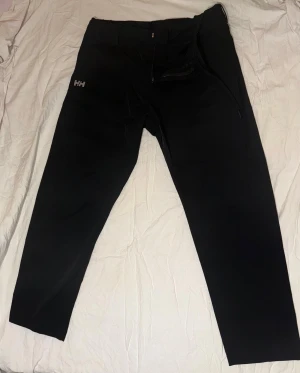 Svarta byxor från Helly Hansen - Svarta byxor från Helly Hansen med stilren design och HH-logga på låret. Byxorna har rak passform, dragkedja i midjan och är tillverkade i ett slitstarkt syntetmaterial som passar perfekt för utomhusaktiviteter. Snygga och funktionella för dig som gillar friluftsliv.