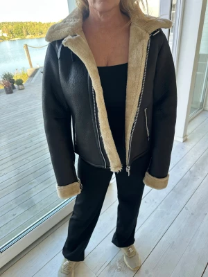 Svart teddyjacka från NA-KD - Säljer en svart teddyjacka från NA-KD i storlek 36. Jackan har beige fuskpälsfoder, bred krage och muddar, samt silvriga dragkedjor framtill och vid ärmsluten. Perfekt för kalla dagar när du vill vara både varm och snygg.