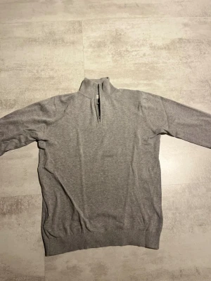 Grå stickad half zip tröja från MNG - Snygg grå tröja från MNG med half zip och hög krage. Tröjan är långärmad och har ribbade muddar vid ärmslut och nederkant. Perfekt för dig som gillar enkel och stilren design. Passar till många olika looks.