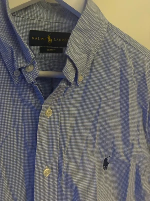 Blårutig slim fit skjorta Ralph Lauren - Snygg blårutig skjorta från Ralph Lauren i slim fit-modell. Skjortan har klassisk krage, knappar framtill och det ikoniska broderade Polo-logot på bröstet. Tillverkad i mjuk bomull och har långa ärmar med manschettknappar. Perfekt för dig som gillar stilren och tidlös design.
