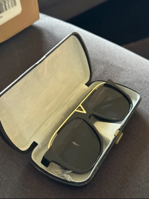 Svarta solglasögon med gulddetaljer - Snygga solglasögon med svart båge och mörka glas. Ramen har coola gulddetaljer och en unik V-formad detalj framtill. Skalmarna är också svarta med guldinslag. Kommer i ett stilrent fodral.