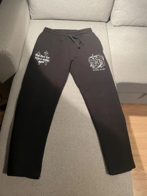 Svarta mjukisbyxor Superior Limited Edition - Svarta mjukisbyxor med tryck i vitt och ljusblått på båda benen. Ena sidan har texten 'New York City Limited Edition USA' med kors, andra sidan har ett stort S och D med stjärnor. Byxorna har snörning i midjan och är riktigt sköna för chill eller streetwear.