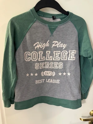 Grå och grön college-tröja strl: 146/152 - College-tröja med grå kropp och gröna ärmar, tryck med texten 'High Play College Series 1973 Best League' framtill. Rund hals och muddar i grönt. Materialet är 65% bomull och 35% polyester, perfekt för chill dagar.