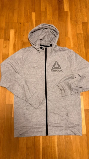Grå zip-hoodie från Reebok, stl S - Ljusgrå hoodie från Reebok med svart dragkedja och justerbar huva. Jackan har Reebok-logga på bröstet, långa ärmar och är tillverkad i mjuk bomullsblandning. Perfekt för chill eller träning.