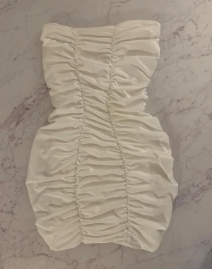 Vit axelbandslös kortklänning - Supersnygg vit axelbandslös kortklänning med cool rynkad design framtill och baktill. Klänningen har en figurnära passform och är perfekt för dig som vill sticka ut med en modern och trendig look. Materialet känns mjukt och har en lätt glans.