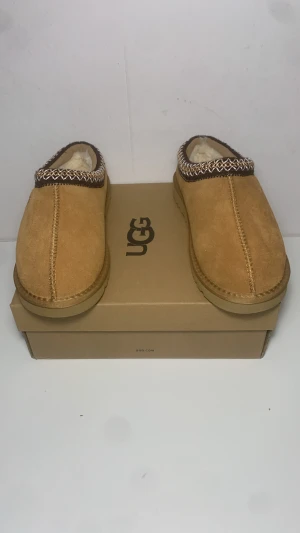 UGG slip-in mockar - Säljer ett par UGG slip-in mockasiner i beige mocka med fluffigt foder och snyggt broderad kant i brunt och vitt. Skorna har rund tå och platt sula med grovt mönster undertill för extra grepp. Perfekta för chill dagar och håller fötterna varma.