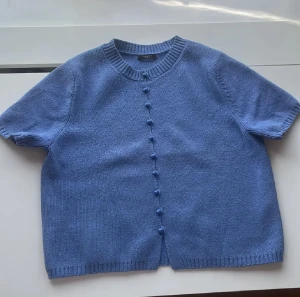 Blå stickad kofta från Massimo Dutti - Supersöt blå stickad kofta från Massimo Dutti med korta ärmar och rund hals. Koftan har matchande blå knappar framtill. Varan är i nytt skick och handlad på Massimo Duttis egna butik i Stockholm. 