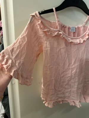 Ljusrosa volangblus från Lindex strl: 134 - Supersöt ljusrosa blus från Lindex med volangdetaljer vid halsringning, ärmslut och nederkant. Blusen har offshoulder-modell med smala axelband och är tillverkad i mjuk, krinklad viskos som ger en luftig känsla. Perfekt för dig som gillar romantisk och trendig stil.