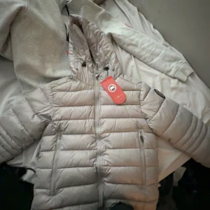 Ljusgrå dunjacka från Canada Goose  - Säljer en snygg ljusgrå dunjacka från Canada Goose med huva och dragkedja framtill. Jackan har två dragkedjeförsedda fickor, broderad logotyp på ärmen och quiltad design. Perfekt för kalla dagar och riktigt stilren look.