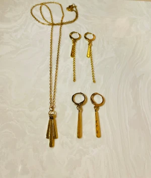 Set med guldiga örhängen och halsband - Snyggt set med två par guldiga örhängen och matchande halsband. Örhängena har runda hoops med hängande stavar, ett par med kedja och ett par utan. Halsbandet har tre smala, hamrade hängen. Perfekt för dig som gillar stilrena och trendiga accessoarer.