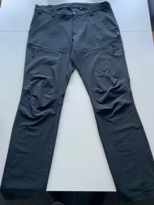 Trekmates byxor  - Svarta friluftsbyxor från Trekmates i storlek XL, med raka ben och praktiska dragkedjor vid fickorna. Byxorna har bälteshällor och är gjorda i ett slitstarkt syntetmaterial som passar perfekt för utomhusaktiviteter. Snygg detalj med logotypen på benet.