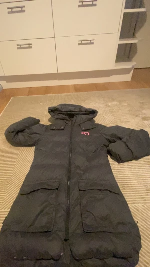 Svart lång dunjacka Kari Traa XS - Lång svart dunjacka från Kari Traa i storlek XS. Jackan har huva, stora fickor med lock, dragkedja framtill och coola rosa detaljer. Perfekt för kalla dagar och har extra dragkedjor i sidorna för mer rörelsefrihet.