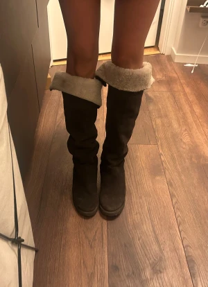 Bruna höga mockastövlar med fårfoder - Säljer ett par bruna höga stövlar i mocka med mjukt, ljust fårfoder som kan vikas ner för en cool look. Stövlarna har platt sula och rund tå, perfekta för kalla dagar. De är klassiska och passar till många outfits.