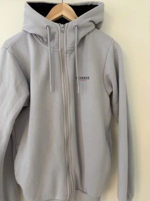 Ljusgrå hoodie från McKenzie - Säljer en ljusgrå hoodie från McKenzie med dragkedja och huva med snörning. Tröjan har ribbade muddar och lång ärm, perfekt för chill dagar. Materialet är mjuk bomull och polyester, med diskret logga på bröstet. Snygg och enkel stil som funkar året runt. Byxor finns också med och de kostar 150 kr.