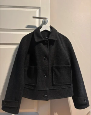Svart ulljacka från 157, storlek S - Stilren svart jacka från 157 i ullmix med klassisk krage, stora framfickor och knäppning med knappar. Jackan har en boxig, något croppad passform och långa ärmar med knappdetalj vid ärmslut. Perfekt för höst och vinter.