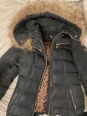 Svart pufferjacka från Hollies - Säljer en snygg svart pufferjacka från Hollies med äkta päls på huvan. Jackan är väldigt använd där efter priset 