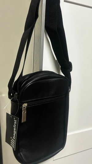 Svart axelväska från Bagsfirst - Stilren svart axelväska från Bagsfirst i konstläder med justerbar rem och dragkedja upptill. Väskan har ett huvudfack med innerficka och är perfekt för att bära det viktigaste. Enkel och clean design som passar till många outfits.