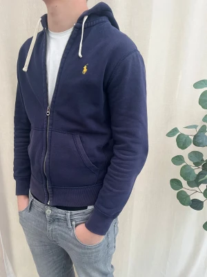 Marinblå hoodie från Polo Ralph Lauren - Snygg marinblå hoodie från Polo Ralph Lauren med gul broderad logga på bröstet. Tröjan har huva med vita snören, dragkedja framtill och två fickor. Lite för liten för mig nu därför jag säljer den