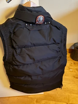 Svart dunväst från Parajumpers - Snygg svart dunväst från Parajumpers med hög krage, dragkedja och flera fickor framtill. Västen har coola detaljer som patch på ryggen och orange inslag vid axeln. Perfekt för lager-på-lager och streetstyle.