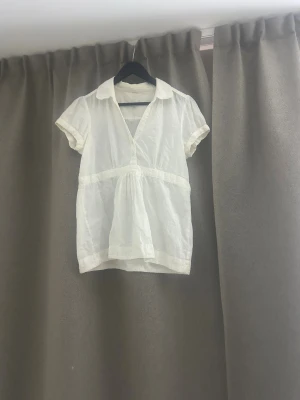 Vit kortärmad blus med spetsdetaljer - Söt vit blus i tunn bomull med korta puffärmar och spetsdetaljer på krage, ärmslut och nederkant. Djup v-ringning med knappar framtill och lätt rynk under bysten för en luftig känsla. Perfekt för en fräsch och somrig look.