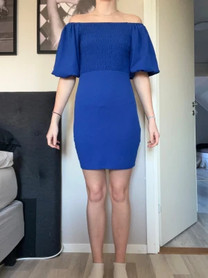 Blå offshoulder kortklänning - Säljer en snygg blå offshoulder klänning med smockad överdel och vida, korta ärmar. Klänningen är figurnära och slutar ovanför knäna. Perfekt för dig som vill ha en stilren och trendig look med lite volym på ärmarna.