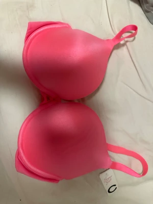 Rosa push up BH D75 Super Soft - Säljer en snygg push up BH i stark rosa färg, modell U Stacey Push up. Mjuka, vadderade kupor och justerbara axelband. Materialet är slätt och känns supermjukt mot huden. Perfekt för dig som vill ha extra lyft och en poppig färg i underklädeslådan.