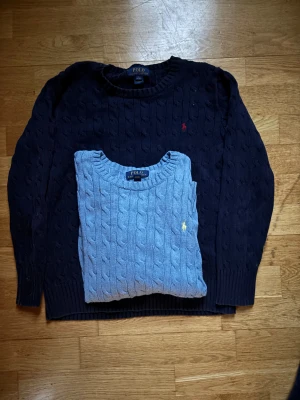Marinblå och ljusblå stickad tröja Polo - Två klassiska stickade tröjor från Polo Ralph Lauren, en i marinblått med röd broderad logga och en i ljusblått med gul broderad logga. Båda har kabelstickat mönster, rund hals och ribbade muddar. Den ljusblåa har en liiiten deffekt under armen som inte syns eller märks vid användning, Hör av er vid behov av bild! 