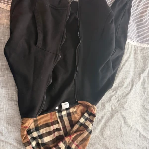 Svart Burberry hoodiejacka med rutig huva - Svart hoodiejacka från Burberry med dragkedja och stor huva fodrad i klassiskt beige, svart och röd rutig design. Jackan har två fickor framtill och är tillverkad i mjukt bomullsmaterial. Perfekt för dig som vill ha en stilren men ändå ikonisk look.