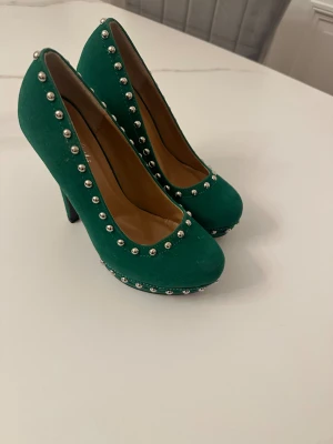 Gröna pumps med nitar och hög klack - Snygga gröna pumps från Milafish i mockaimitation med rund tå och hög klack. Skorna har silverfärgade nitar längs kanterna och runt sulan för en edgy look. Perfekta för dig som vill sticka ut med färg och detaljer.