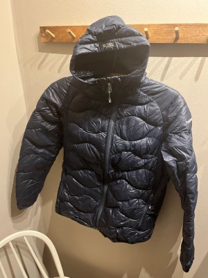 Peak Performance: Mörkblå dunjacka - Snygg mörkblå dunjacka från Peak Performance med quiltad design och huva. Jackan har dragkedja framtill och två sidofickor med dragkedja. Ett pyttigt hål fram (se bild, därav pris), i övrigt i bra skick. 