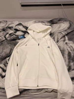 Polo kofta - Vit hoodie från Polo Ralph Lauren med dragkedja och snörning i huvan. Klassisk svart broderad logga på bröstet. Mjuk bomullskvalitet, två fickor fram och långärmad modell. Perfekt för chill dagar och enkel att matcha med jeans eller joggers.