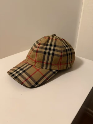 Burberry rutig keps beige M - Säljer en klassisk Burberry keps i beige med det ikoniska rutmönstret i svart, vitt och rött. Kepsen har böjd skärm, justerbart spänne i silver med Burberry-logga och är tillverkad i bomull. Perfekt accessoar för en trendig och tidlös look.