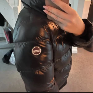 Colmar jacka glansig - Säljer en svart, glansig dunjacka från Colmar med hög krage och dragkedja framtill. Jackan har quiltad design och Colmar-logga på ärmen. Perfekt för kalla dagar när du vill hålla stilen och värmen.. Den är väldigt varm och bra för vintern. Jag är 168cm och brukar ha på mig storlek S men den passar bra på mig också🤍