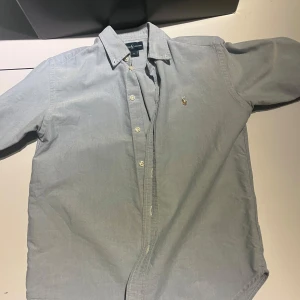 Ljusblå skjorta från Ralph Lauren - Snygg ljusblå skjorta från Ralph Lauren med klassisk krage och knappar framtill. Skjortan har det ikoniska broderade Polo-logot på bröstet i guld och blått. Tillverkad i mjuk bomull, perfekt för en clean och stilren look.