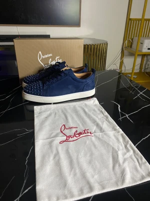 Blå Christian Louboutin sneakers med nitar - Säljer ett par mörkblå sneakers från Christian Louboutin i mocka med coola nitar på tån och klassisk röd sula. Skorna har snörning och vit platt sula, samt en lyxig vibe som sticker ut. Perfekt för dig som gillar statement pieces och exklusiva detaljer.