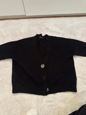 Svart ribbad kofta från LCW Casual - Svart stickad kofta från LCW Casual med ribbad struktur och tre stora knappar framtill. Modellen har v-ringad hals och långa ärmar, perfekt för lager-på-lager. Materialet är mjukt och ger en chill vibe till din outfit.