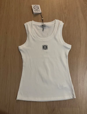 Vit ribbad linnetopp från Loewe - Säljers en fräsch vit ribbad linnetopp från Loewe med broderad svart logga framtill. Toppen är ärmlös och har en rundad halsringning. Materialet är mjuk bomull som sitter skönt mot kroppen och ger en clean och stilren look.