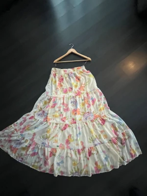 Blommig långkjol från Boohoo - Superfin långkjol från Boohoo med volanger och blommigt mönster i rosa, gul, lila och grönt på vit botten. Kjolen har resår i midjan och är gjord i ett lätt, flowy material som känns perfekt för varma dagar.  Säljer på grund av att den är för stor men priset kan diskuteras!☺️