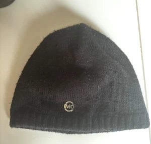 Svart stickad mössa från Michael Kors - Snygg svart stickad mössa från Michael Kors med diskret MK-logga i metall framtill. Klassisk form och ribbad kant nedtill. Perfekt accessoar för kyliga dagar och enkel att matcha med olika outfits. Liten i storlek 