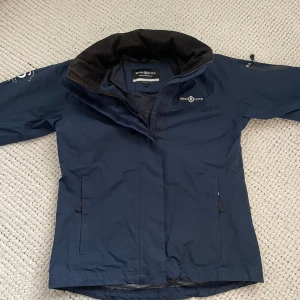 Marinblå vindjacka från Henri Lloyd - Snygg marinblå vindjacka från Henri Lloyd med hög krage, avtagbar huva och dragkedja framtill. Jackan har praktiska fickor med dragkedja och diskret logga på bröstet. Perfekt för blåsiga dagar och stilren look. Pris kan självklart diskuteras. Nypris 1400