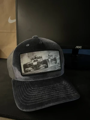 Svart keps med F1-motiv - Cool svart keps med mesh på sidorna och ett stort tryck framtill med Formel 1-bilar i svartvitt. Kepsen är tillverkad i 100% polyester och har en klassisk böjd skärm. Perfekt för dig som gillar motorsport och vill sticka ut med en unik detalj.