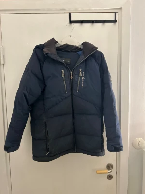 Blå Sail Antarctica dunjacka herr - Min vän säljer en mörkblå dunjacka från Sail Antarctica med huva, två bröstfickor med dragkedja och patch på ärmen. Jackan har quiltad design och är tillverkad i syntetmaterial. Perfekt för kalla vinterdagar och har en sportig vibe. Jackan är i stolek 160. Är lite sönder på baksidan men är sydd. Den tvättas innan den skickas.