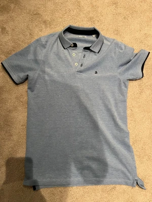 Blå pikétröja från Jack & Jones - Snygg ljusblå pikétröja från Jack & Jones med klassisk krage och tre knappar framtill. Tröjan har svarta detaljer på krage och ärmslut samt ett litet broderat S på bröstet. Perfekt för dig som gillar stilren och enkel design.