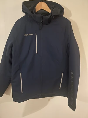 Mörkblå Bauer vinterjacka med huva - Säljer en ny mörkblå Bauer vinterjacka i storlek M. Jackan har huva, tre dragkedjeförsedda fickor och snygga vita detaljer vid dragkedjorna. Perfekt för kalla dagar och sportiga outfits. Bauer-logga på bröstet och ärmen för en sportig look. 