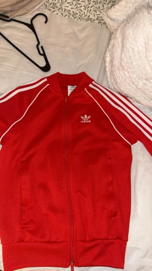 Röd Adidas track jacket med vita ränder - Röd Adidas tröja med klassiska vita ränder längs ärmarna och vit logga på bröstet. tröja har dragkedja framtill, ribbade muddar och sportig look. Perfekt för dig som gillar streetstyle och sportiga vibes.