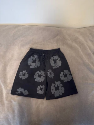 Denim tears shorts - Säljer ett par svarta shorts från Denim Tears med gråa blommönster över hela tyget. De har elastisk midja med snörning och en bakficka. Materialet känns som polyester, perfekt för chill dagar. Snygg och unik design som sticker ut.PRIS KAN DISKUTERAS